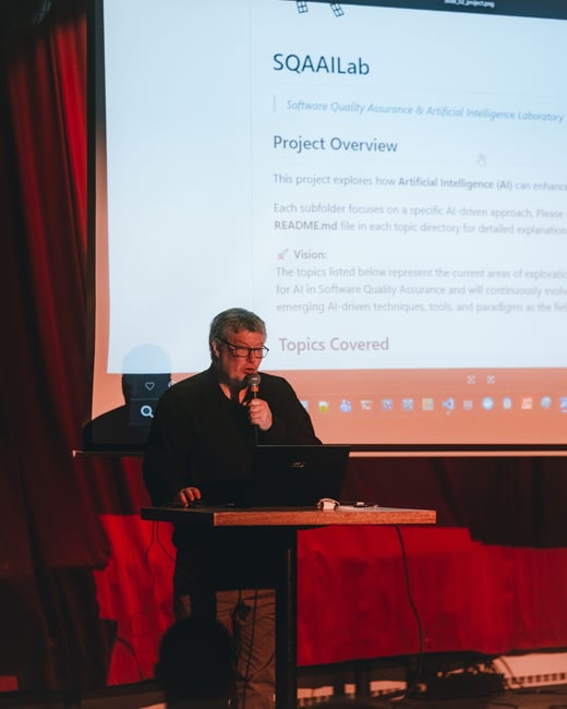 A man is giving a presentation on a stage, standing behind a laptop and speaking into a microphone with a large projection screen displaying project details behind him.