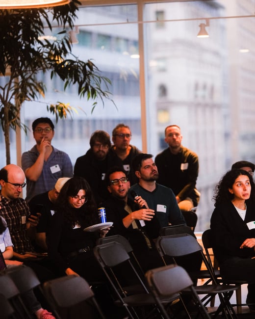 A candid photograph of a group of people attending an indoor event, with one man in the center speaking into a microphone while others listen.
