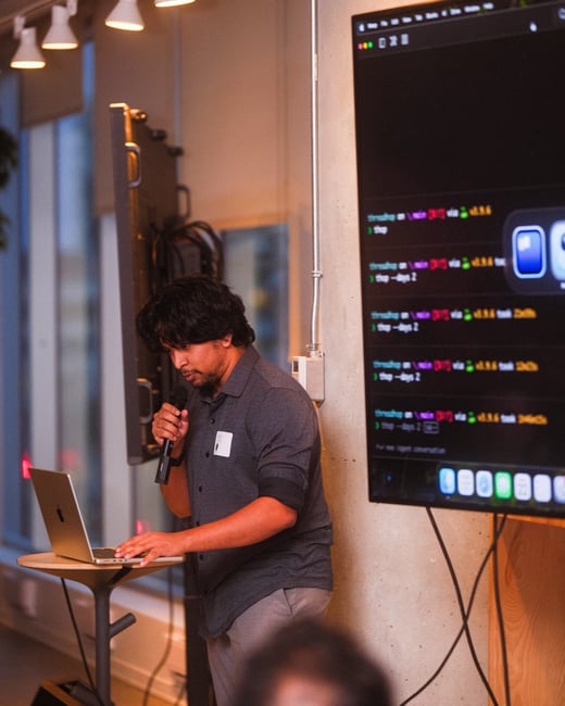 A man is giving a presentation, holding a microphone and looking at a laptop, with a large screen behind him displaying terminal commands and code.