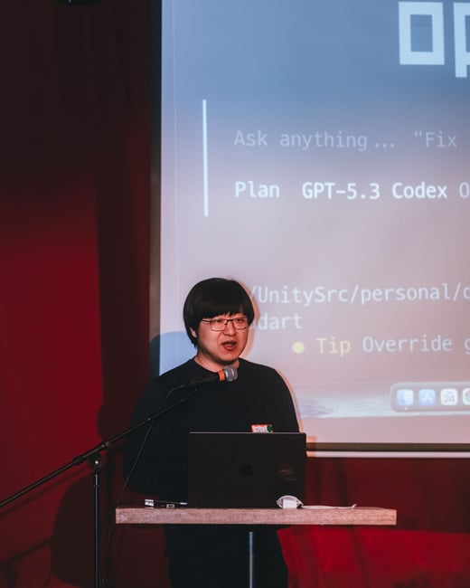 A man is giving a presentation on a stage, standing behind a podium with a laptop and a microphone, with a large screen behind him displaying a code editor interface.