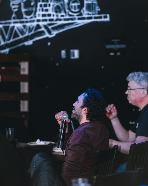 A man with curly hair holds a microphone while seated at a table, participating in a discussion. Behind him is a dark wall featuring a large white line drawing of what appears to be a brewery or industrial setup.