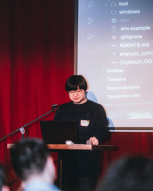 A man is giving a presentation on a stage, standing behind a podium with a laptop while a screen behind him displays a code editor's file structure.