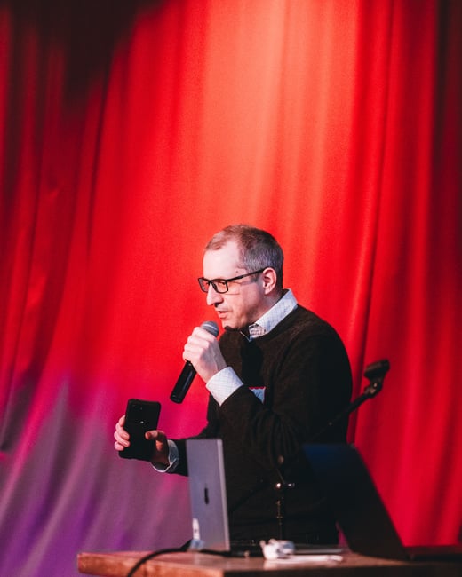A man wearing glasses and a dark sweater speaks into a microphone while holding a smartphone, standing behind a table with laptops in front of a vibrant red curtain.