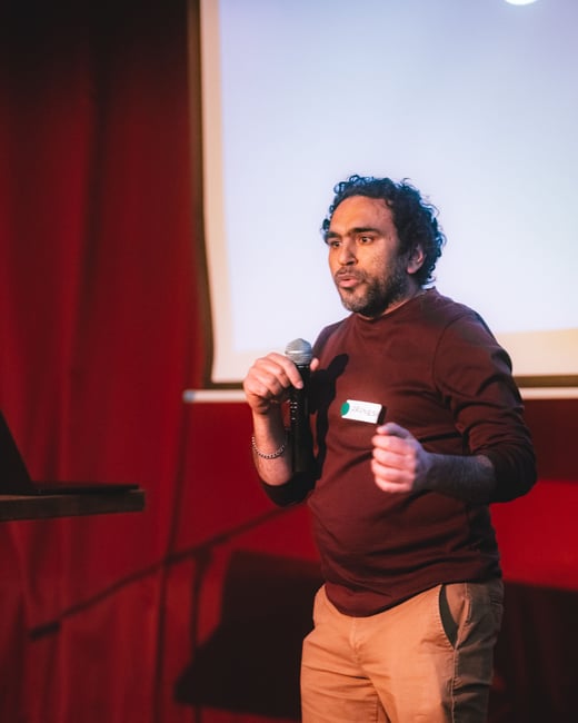 A man stands on a stage, holding a microphone and gesturing while giving a presentation. He is wearing a maroon shirt and tan trousers, with a name tag visible on his chest.