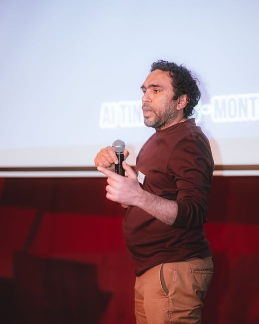 A man is standing on a stage, holding a microphone and speaking during a presentation in front of a projection screen.