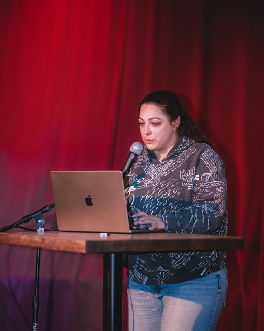 A woman stands behind a high table, using a laptop and speaking into a microphone against a backdrop of a red curtain.