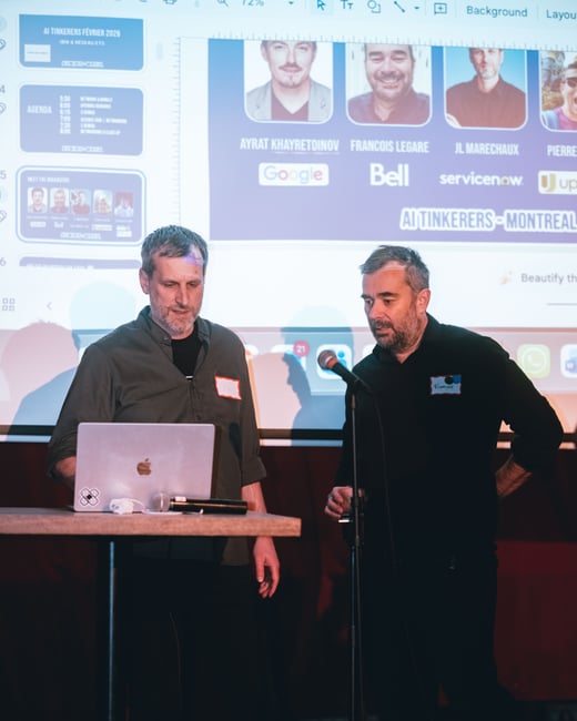 Two men are on a stage during an event, with one looking at a laptop on a high table and the other standing near a microphone. A large projection screen behind them displays an agenda and speaker profiles for an 'AI Tinkerers' event in Montreal.