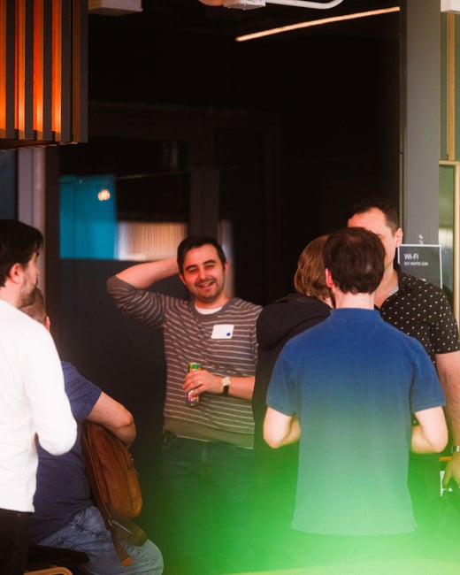 A group of people are socializing in an indoor office or event space, with one man in a striped sweater smiling toward the camera.