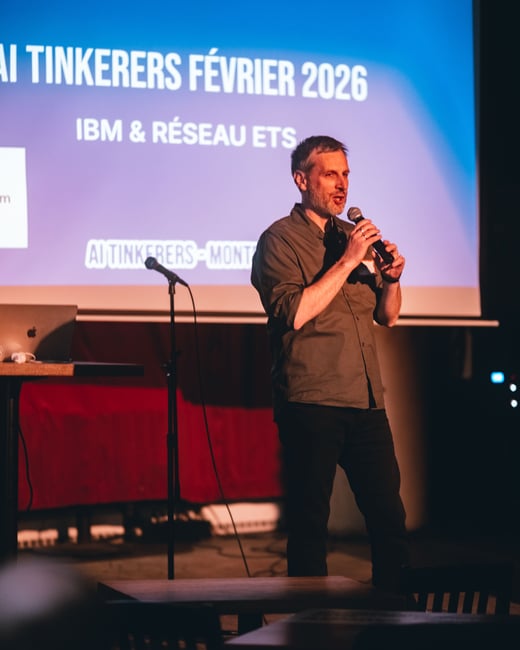A man stands on a stage, speaking into a handheld microphone in front of a large projection screen during an event.