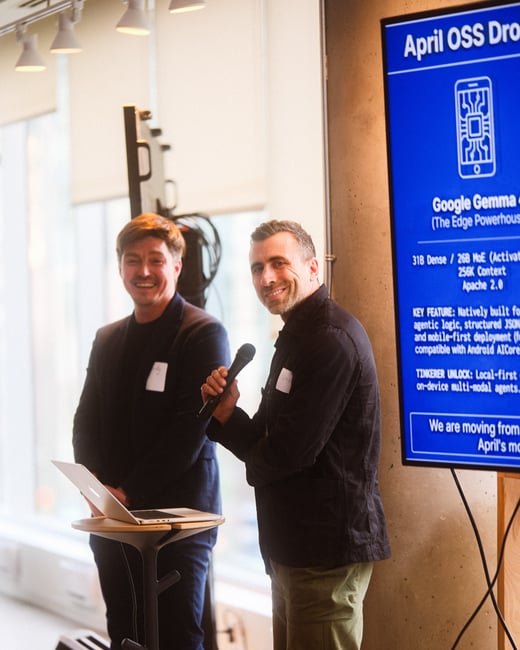 Two men are giving a presentation in a modern indoor space. One man holds a microphone and smiles at the camera, while the other stands behind a laptop on a tall stand.