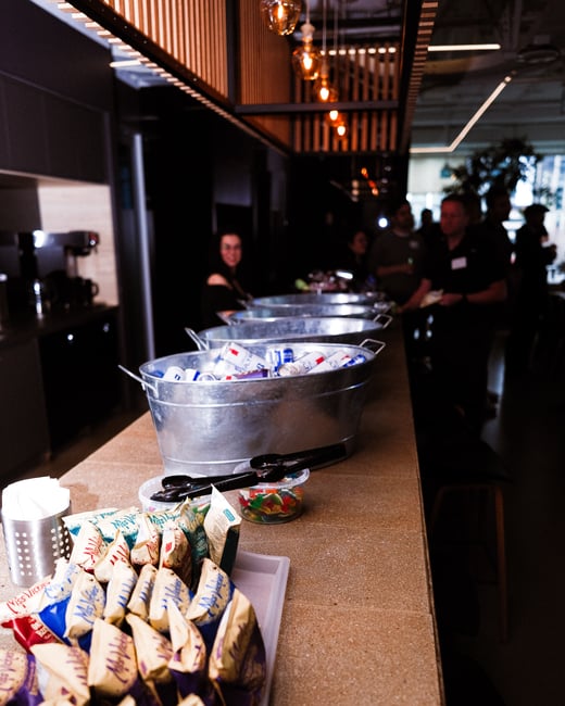 A long counter at an event is stocked with various bags of Miss Vickie's chips and large metal tubs filled with canned beverages. In the background, a group of people are socializing in a modern, dimly lit indoor environment.