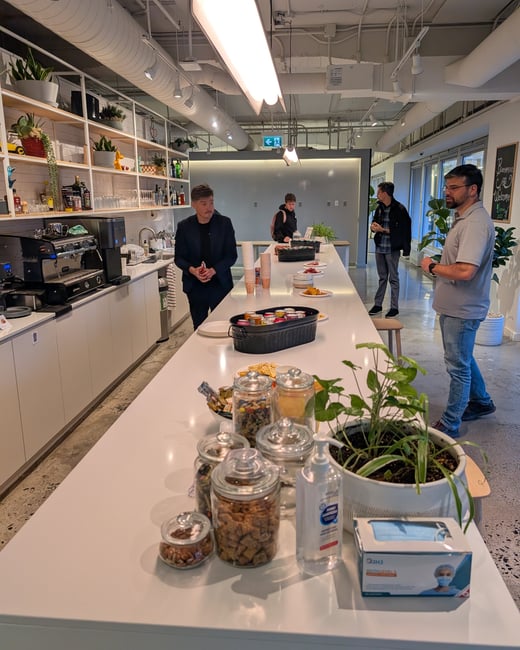A candid photograph taken inside a modern office break room or kitchen area, showing a long white counter filled with snacks, drinks, and supplies, where four men are casually gathered.