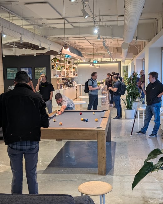 A candid photo showing a group of men socializing and playing pool in a modern, industrial-style office common area that features high ceilings, concrete floors, and a brightly lit kitchenette/coffee bar.