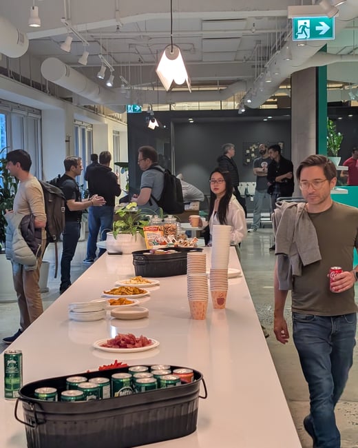 A candid photograph capturing a social gathering or break in a modern office, showing several people mingling around a long white table laden with snacks, stacks of cups, and a tub of canned drinks.