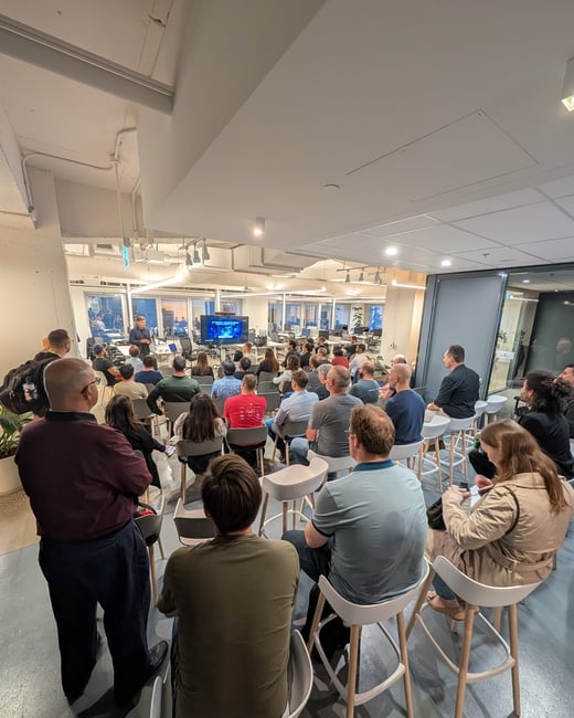 A photograph capturing a large audience seated in a modern office space, watching a presenter standing near a screen at the front of the room during an event or talk.