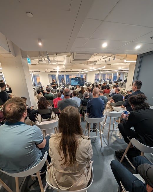 A photograph taken from the back of an audience showing a large group of people seated on high stools, attending a presentation in a modern, brightly lit office space during the evening. A speaker stands next to a large screen displaying the title 'What's new in AI?'.