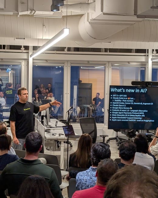 A photograph captures a man giving a presentation on 'What's new in AI?' to a large seated audience in a modern, industrial-style indoor venue with exposed ductwork.