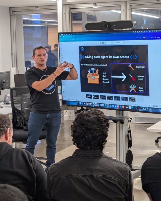 A man is giving a technical presentation in an office setting, standing next to a large display screen that shows a slide about agent access and tools in software development.
