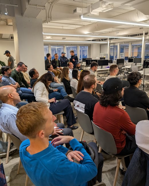 A candid photograph showing a large group of people seated in a modern, industrial-style office space, attending a presentation or meeting. The audience is primarily facing away from the camera toward the front of the room.