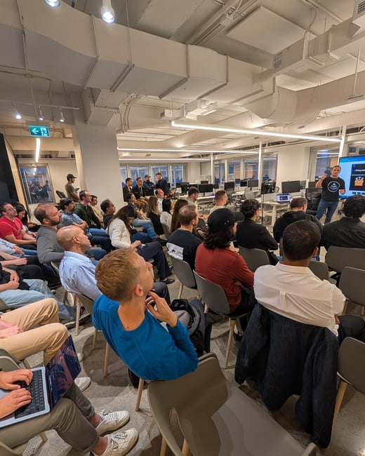 A candid photograph taken from within an audience, showing a large group of people seated in an indoor office space attending a presentation given by a speaker standing near a projection screen.