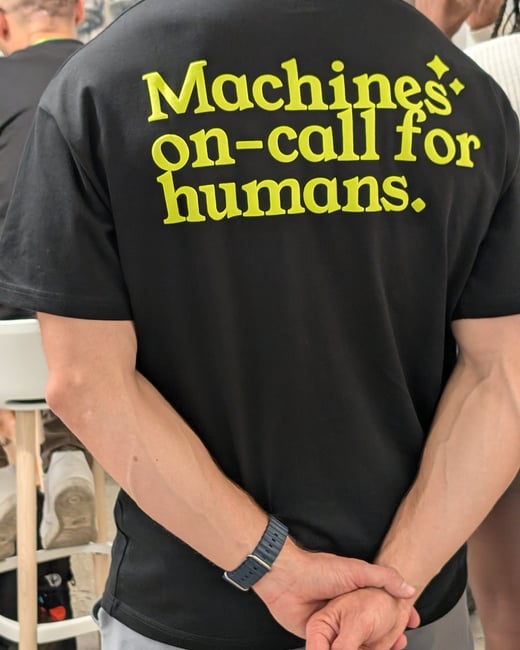 A close-up photograph of a person's back wearing a black T-shirt that features the bright yellow slogan, "Machines' on-call for humans."