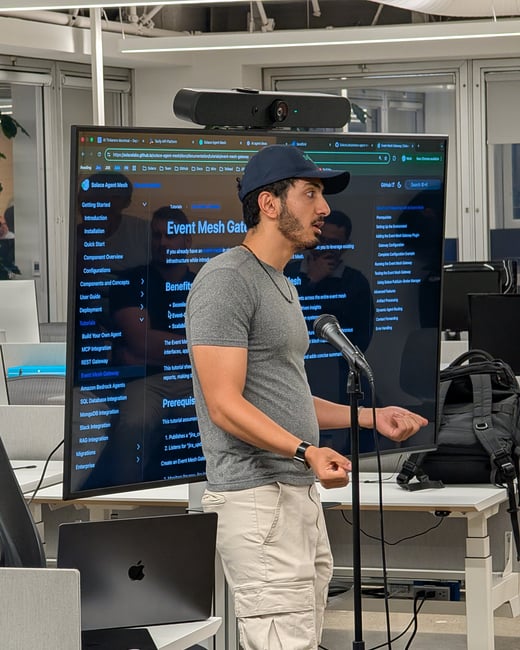A photograph showing a man giving a technical presentation or talk in a modern office setting, standing in front of a large monitor displaying documentation related to 'Solace Agent Mesh' and 'Event Mesh Gateway'.