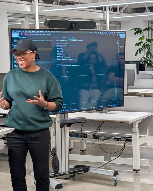 A woman wearing a dark green sweater and a baseball cap is giving a presentation in a modern office, standing next to a large monitor displaying software code. Reflections of audience members are visible on the screen.