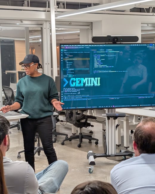 A woman wearing a dark green sweatshirt and a baseball cap is giving a technical presentation or demo in a modern office setting, standing next to a large monitor displaying code and the word 'GEMINI'. Several attendees are visible in the foreground, watching the presentation.