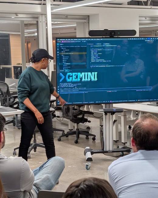 A woman wearing a dark green sweater and a baseball cap is giving a technical presentation in an office environment, pointing at a large monitor displaying code and the prominent pixelated word "GEMINI". Several audience members are seated in the foreground, facing the screen.