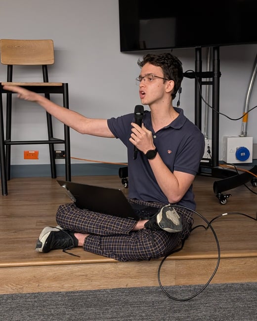A young man wearing glasses and a navy polo shirt is sitting cross-legged on a raised wooden platform, holding a microphone and gesturing widely while giving a presentation. He has a laptop resting on his lap, set against a background featuring a gray wall, a high stool, and a large black screen.