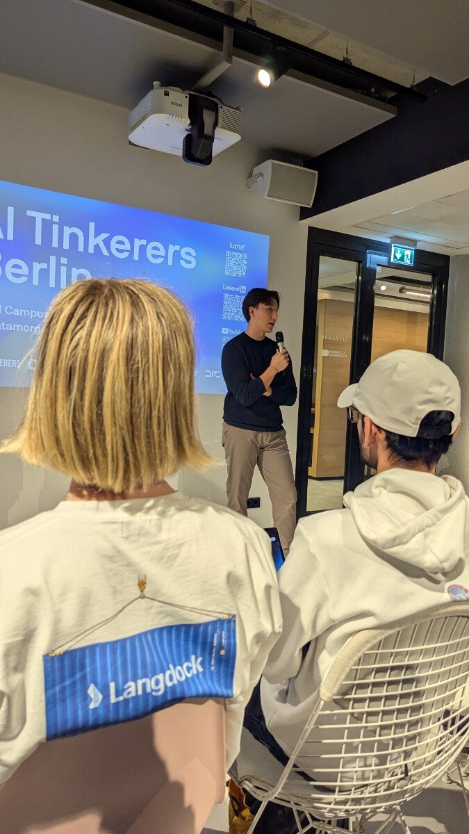 A photograph capturing an indoor presentation or tech talk, showing a male speaker holding a microphone in front of a projected screen displaying the text "AI Tinkerers Berlin." Two audience members, whose backs are visible in the foreground, are seated, one wearing a white shirt featuring the "Langdock" logo.