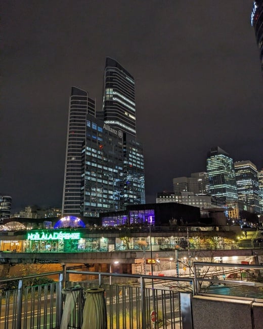 A nighttime photograph showcasing the illuminated skyscrapers of a modern business district, likely La Défense in Paris, viewed from an elevated perspective over a railing and a lower commercial structure featuring bright green neon signage.