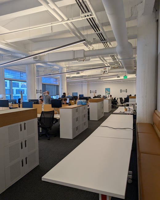 A modern, open-plan office space featuring long white desks, people working at computer stations, and an industrial-style ceiling with exposed white pipes and ductwork.