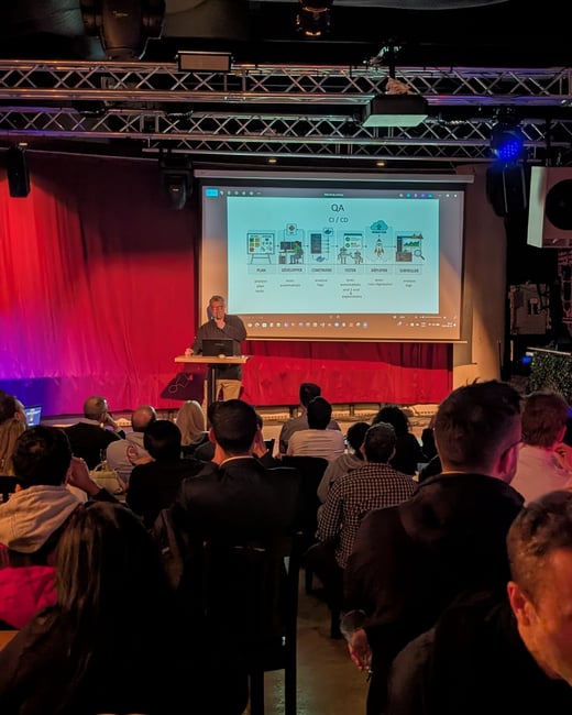 A man stands at a podium giving a technical presentation on a stage with a projector screen showing a QA and CI/CD workflow diagram to a seated audience.