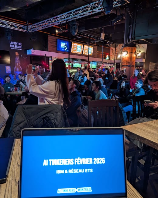 A photograph taken from a first-person perspective at a tech meetup, showing a laptop screen in the foreground and a crowded bar setting in the background.