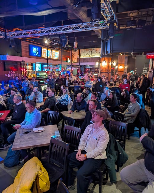 A large audience is seated in a dimly lit bar or lounge, attentively watching a presentation or event. The venue features industrial-style ceilings with stage lighting and a bar area in the background.
