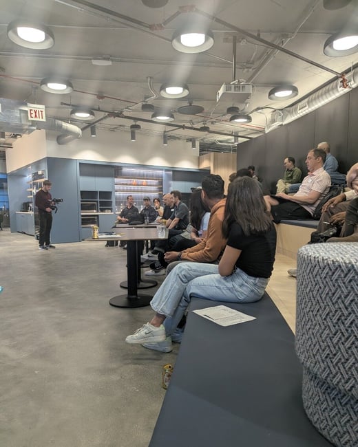 An indoor photograph showing an audience seated on tiered benches and at tables in a modern office or event space, listening to a presentation while a videographer stands to the left.