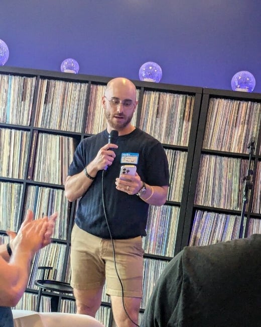 A man stands in front of a large shelf filled with vinyl records, speaking into a microphone while looking at his smartphone.