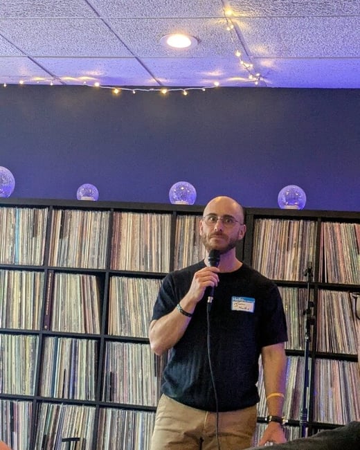 A man wearing glasses and a black t-shirt stands holding a microphone, speaking in front of a large bookshelf filled with vinyl records.