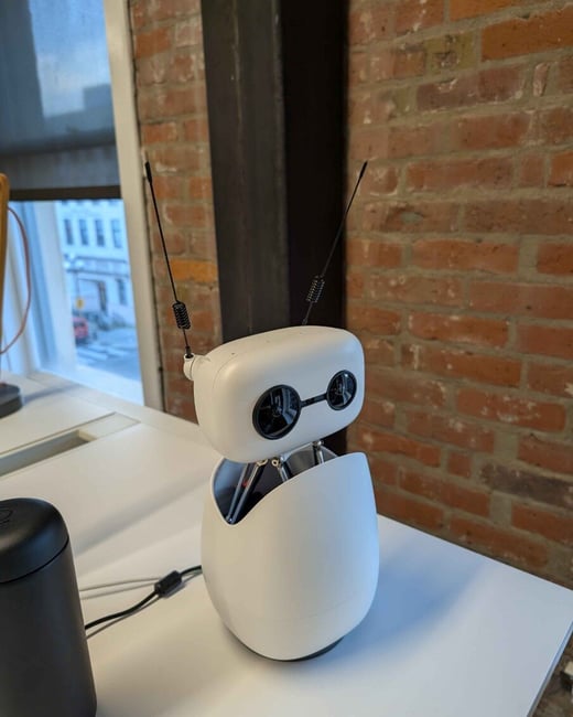 A small, white, rounded robot with two large black eyes and thin antennae sits on a white desk in an indoor setting with a brick wall background.