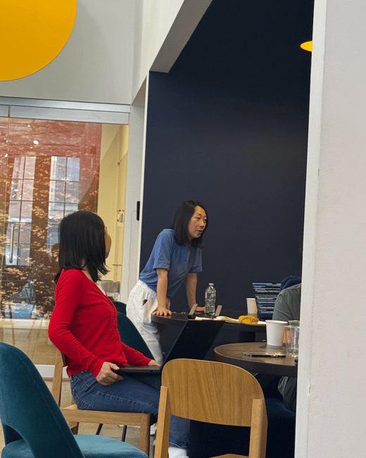 A candid photograph capturing a small group meeting indoors in a modern office space, featuring two women, one seated with a laptop and another standing and speaking at a dark table.