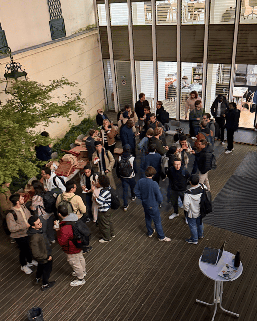 A high-angle photograph captures a large group of people socializing and eating pizza in an outdoor courtyard area next to a modern building with large glass windows during the evening or night.