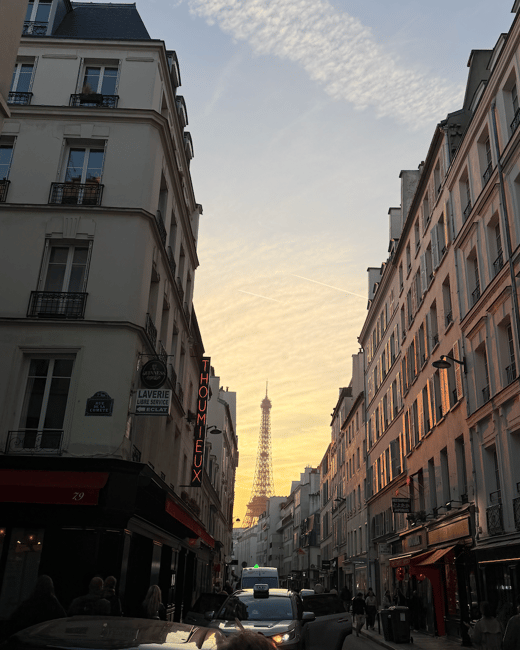 A photograph capturing a narrow Parisian street at sunset, where classic Haussmann buildings frame a view of the Eiffel Tower silhouetted against a vibrant golden sky. The street below is busy with cars and pedestrians, highlighted by the warm evening light reflecting off the right-side buildings.