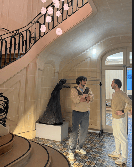 Two men are standing and engaged in conversation in a grand, ornate interior space featuring a winding staircase, patterned tile floor, and a modern chandelier composed of numerous glowing geometric shapes.
