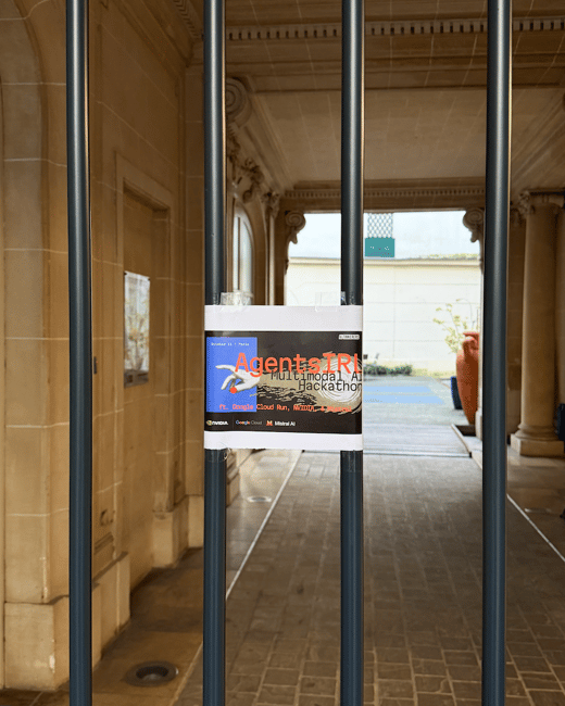 A photograph showing a small event poster taped to the vertical bars of a dark metal gate, set against the backdrop of a light-colored stone building entryway leading to a courtyard.