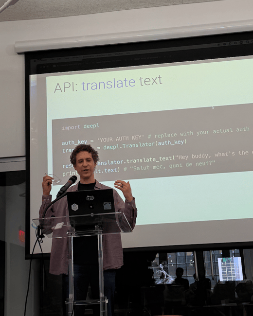 A man stands behind an acrylic podium presenting technical code (Python/Deepl API) projected onto a large screen in a dimly lit indoor venue, addressing an audience visible in the foreground shadows.