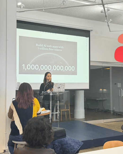 A photograph captures a woman giving a presentation on a small stage in a modern indoor venue, addressing an audience visible in the foreground. The large projection screen behind her displays text related to building AI web apps and a quantity of 1 trillion tokens.