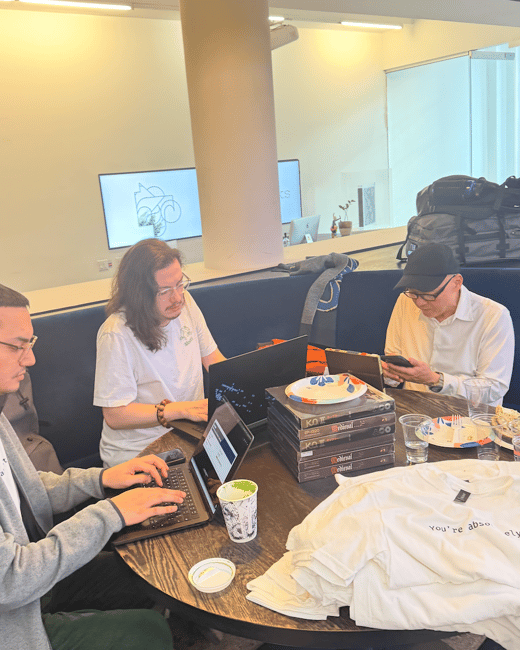 A candid indoor photograph capturing three individuals seated around a circular table in a modern office or co-working space, two of whom are actively working on laptops while the third checks his phone.