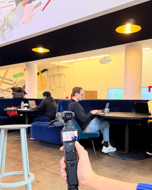 A close-up photograph shows a hand holding a gimbal camera (DJI Osmo Pocket) in a modern office or co-working lounge, while people work on laptops in the background. The scene features blue seating, dark tables, and a large, colorful abstract mural on the ceiling/upper wall.
