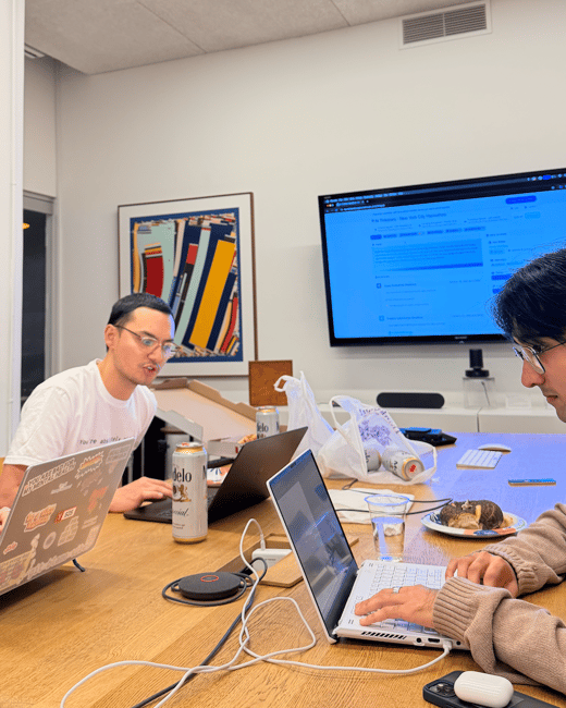 A candid photograph captures two individuals working on laptops at a large wooden table in a modern office or meeting room, with food, drinks (including Modelo beer), and various cables spread across the surface.